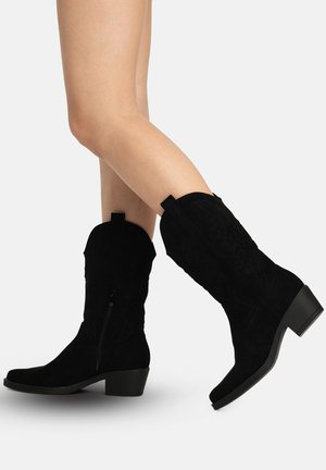 Person iført sorte midt-lægs ruskind cowboystøvler med dekorative syninger og lave blokhæle på en hvid baggrund.