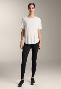 Tricou alb cu mâneci scurte și hem rotund, asortat cu leggings negri strâmți și adidași negri, pe un fundal neutru.