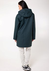 Manteau imperméable vert foncé avec capuche, design mi-long, associé à un pantalon de survêtement gris clair et des baskets noires. Texture lisse, style simple.