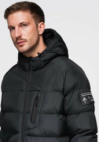 Schwarze Steppjacke mit hohem Kragen, durchgehendem Reißverschluss und Brusttasche. Mit strukturierter Oberfläche und weißen Akzenten an den Beschlägen.