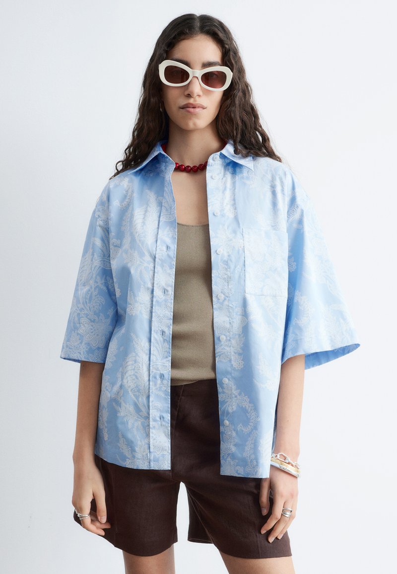 Femme portant une chemise ample bleu clair à motifs sur un débardeur beige, un short marron foncé, des lunettes de soleil blanches, un collier de perles rouges et des bracelets.