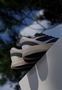 Pair of white, blue, and gray sneakers resting on the edge of a white surface with a blurred background of trees and sky.
