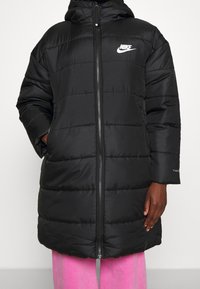 Abrigo acolchado negro con cierre de cremallera, capucha y bolsillos laterales. Presenta costuras horizontales y un logo de Nike en blanco en el pecho.