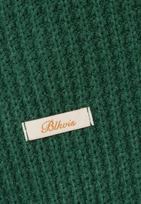 Grüner Strickstoff mit einem eingenähten, cremefarbenen rechteckigen Etikett, auf dem in orangefarbener Schreibschrift "Blkvis" steht.