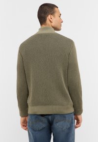 Olivgrüner, gerippter Strickpullover mit Stehkragen, verfügt über eine strukturierte Oberfläche und einen taillierten Saum. Getragen zu blauen Jeans.