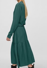 Pitkähihainen, turkoosi mekko, joka on valmistettu sileästä kankaasta; siinä on rypistetty vyötärö ja levenevä alaosa, jossa on kerroksellinen helma. Sopii rentoon istuvuuteen.