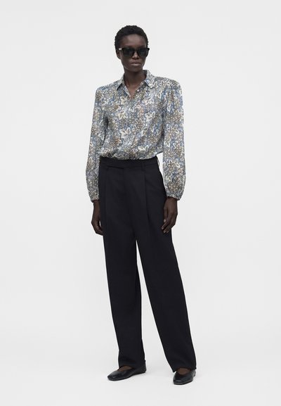Femme portant des lunettes de soleil noires, un chemisier à manches longues à motif floral, un pantalon noir taille haute à jambes larges et des ballerines noires, debout sur fond blanc.