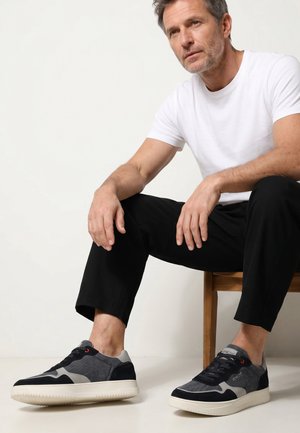 Man van middelbare leeftijd in wit T-shirt en zwarte broek, zittend op een houten kruk, met zwarte en grijze sneakers met witte zolen.