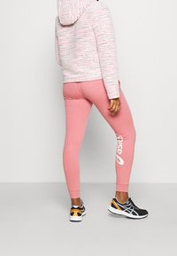 Rosa Sweatpants mit einer enganliegenden Passform, die ein großes weißes Logo an der Seite tragen. Kombiniert mit einem gestreiften Hoodie und schwarzen Sportschuhen.