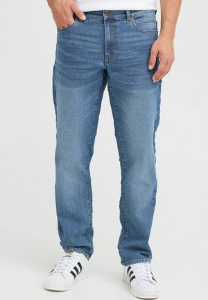 Blaue Denim-Jeans mit geradem Schnitt, ausgestattet mit fünf Taschen, dezenten Verfärbungen sowie einem Knopf- und Reißverschluss, kombiniert mit weißen Sneakers.
