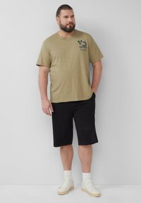 Olijfgroene T-shirt, korte mouwen, grafische print op de borst. Gecombineerd met zwarte shorts en witte sneakers, staand tegen een lichte achtergrond.