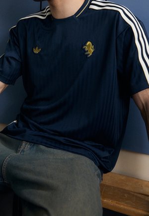 Homme assis sur un banc en bois portant un t-shirt Adidas bleu marine avec des bandes blanches et des logos dorés, associé à un jean bleu.