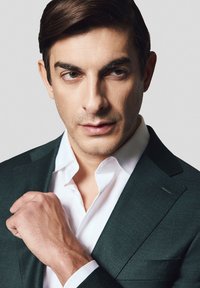 Ciemnozielony żakiet na białej koszuli z wydłużonym kołnierzykiem. Model ma ciemne włosy i poprawia żakiet, trzymając rękę na klapie.