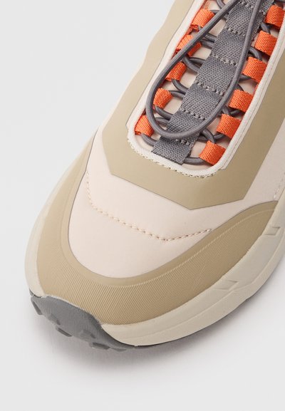 Beige sneaker with a textured rubber outsole, smooth upper, and contrasting orange and gray laces. Features a geometric design on the sides.