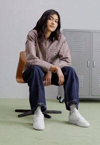 Giacca grigia con colletto e tasca frontale, abbinata a jeans blu larghi e sneakers bianche su un pavimento verde, davanti a uno sfondo chiaro.