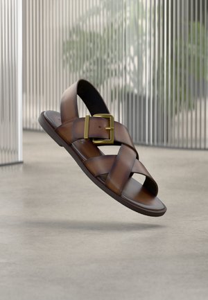 Braune Ledersandale mit breiten, sich kreuzenden Riemen und einer großen goldenen Schnalle, schwebend über einem Betonboden im Innenbereich präsentiert.