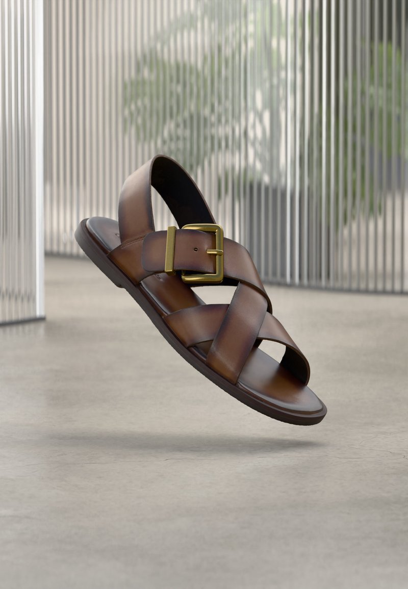 Καφέ δερμάτινο σανδάλι με φαρδιές διασταυρούμενες λουριά και μια μεγάλη χρυσή αγκράφα, εμφανίζεται να αιωρείται πάνω από τσιμεντένιο δάπεδο σε εσωτερικό χώρο.