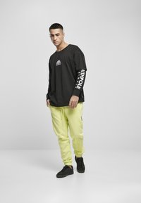 Cămașă neagră cu mânecă lungă, cu grafic alb și siglă pe mâneci, asortată cu pantaloni de trening galbeni neon cu logo. Modelul poartă adidași negri.