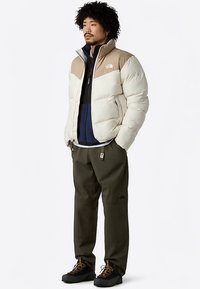 Gewatteerd jack in crème en bruin, met een hoge kraag en ritssluiting. Draagt over een marineblauwe tussenlaag en donkergroene broek, gecombineerd met zwarte laarzen.