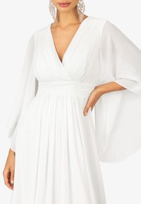 Witte jurk met een V-hals en geplooide bodice, vloeiende mouwen en een ontspannen pasvorm, gemaakt van lichte stof. Verfraaid met ingewikkelde oorbellen.