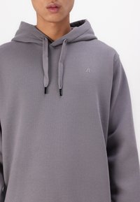 Sudadera con capucha gris hecha de una tela suave, con un bolsillo frontal y un emblema de logo en el pecho. Cuerdas con acentos negros.