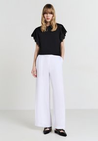 Donna con capelli biondi lisci che indossa un top nero con maniche di pizzo, pantaloni bianchi a gamba larga e scarpe nere a punta con cinturini.