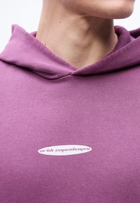 Κοντινό πλάνο ενός ατόμου που φοράει μωβ φούτερ με κουκούλα, με ένα μικρό λευκό λογότυπο «arkk copenhagen» στο κέντρο του στήθους.