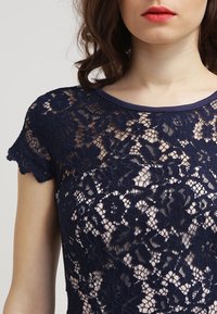 Námořnická krajková blůza s krátkými, vykrojenými rukávy a kulatým výstřihem, zdobená květinovými vzory a poloprůhlednou texturou.