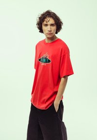 Camiseta de algodón roja con mangas cortas, que presenta un gráfico de llamas en negro y turquesa y el texto "GRMY". Pantalones negros con bolsillos.