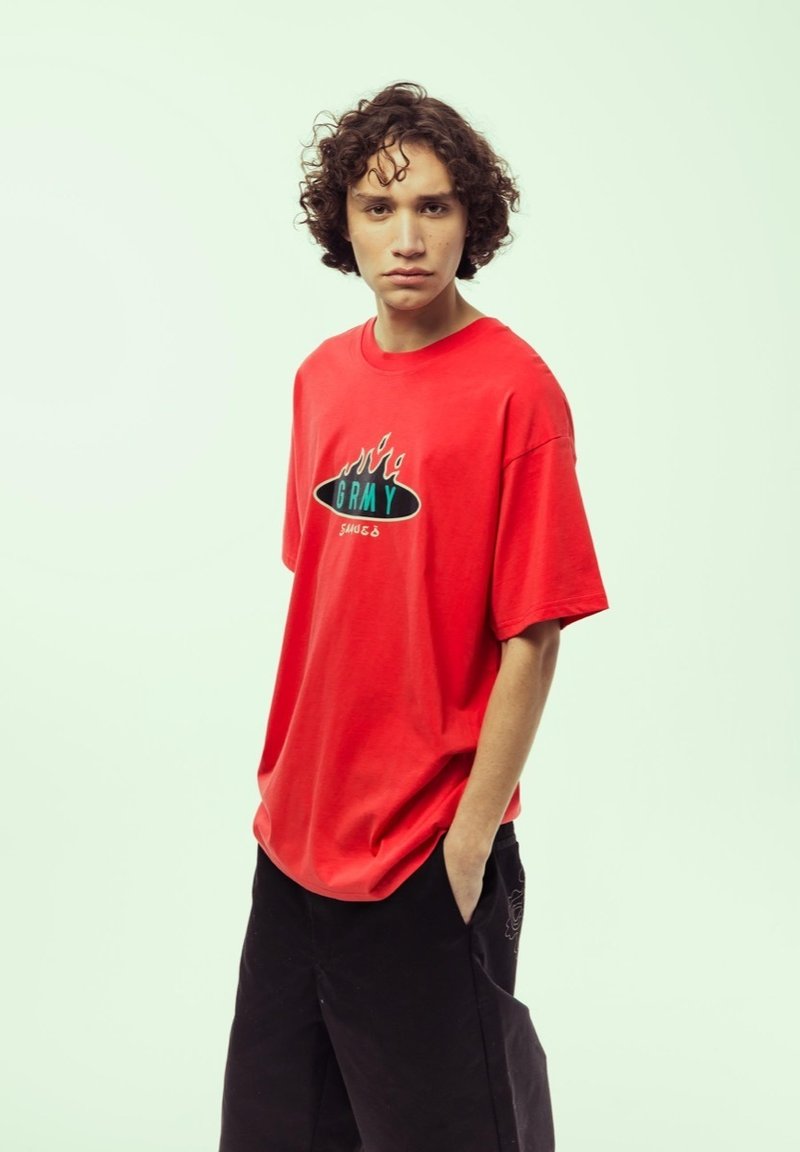Camiseta de algodón roja con mangas cortas, que presenta un gráfico de llamas en negro y turquesa y el texto "GRMY". Pantalones negros con bolsillos.