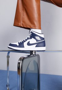 Nike Air Jordan Sneakers aus weißen und marineblauen Leder, hochformatiges Design, mit einem seitlichen Logo und einer weißen Sohle, die auf einer reflektierenden Oberfläche liegen.