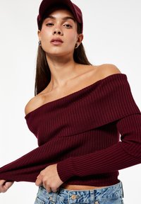 Bordeauxfarbenes, schulterfreies, geripptes Sweatshirt mit langen Ärmeln, das eine horizontale Naht über der Brust hat. Trägt man zu hellen Jeansshorts.