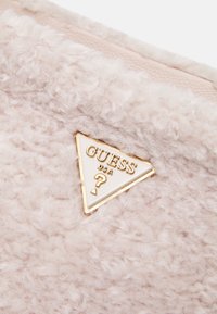 Tessuto in peluche rosa con una texture morbida, caratterizzato da una placca logo triangolare con dettagli dorati che riporta la scritta "GUESS USA."