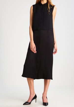 Kobieta w czarnej sukience midi bez rękawów z guzikami z przodu i czarnych szpilkach z ostro zakończonymi noskami, stojąca na neutralnym tle.