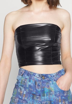 Lähikuva henkilöstä, jolla musta kiiltävä hihaton crop-top ja korkeavyötäröiset siniset farkut abstraktilla silmäkuviolla, käsi taskussa.