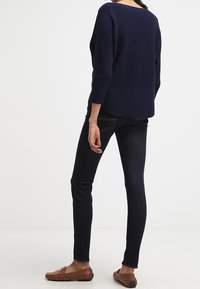 Námornícky modrý žebrovaný sveter, kulatý krk, rukávy 3/4, spárovaný s tmavými úzkými džínami a hnědými mokasínami, pohled zezadu.