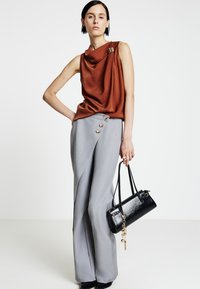 Top marrone senza maniche drappeggiato con accento dorato; pantaloni grigi a gamba larga con dettaglio a bottoni; borsa nera con finiture dorate; texture eleganti.