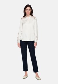 Long-sleeve cream blouse with a front slit and floral embroidery on shoulders, paired with dark denim and light-colored flats.
