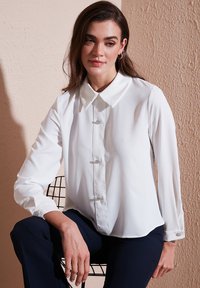 White blouse in lightweight material with long sleeves, a pointed collar, and ornamental button accents on the front and cuffs.