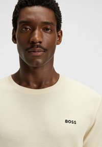 Crèmekleurige, gestructureerde trui met een ronde hals. Heeft een klein zwart "BOSS" logo aan de linkerkant van de borst.