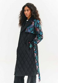Prešívaný čierny trenčkot s kvetinovým vzorom v tyrkysovej a ružovej farbe na golieri a rukávoch, so zviazaným pásom v páse a dvojradovým zapínaním.