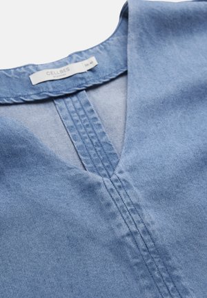 Lyseblå denimbluse med V-hals, synlige sømme langs halsudskæringen og et hvidt "Cellbes" mærke indeni kraven.