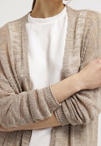 Lightweight, beige knit cardigan layered over a textured white top. Features open front, loose fit, and ribbed cuffs. Subtle pattern detail.