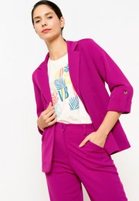 Blazer fucsia su misura con maniche arrotolabili, abbinato a una t-shirt bianca con grafica e pantaloni coordinati; texture liscia e design strutturato.