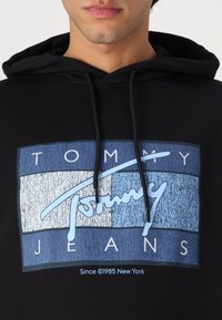 Haut à capuche noir présentant un design graphique bleu et blanc avec le texte "TOMMY JEANS" et des rayures verticales. Détail de cordon sur la capuche.