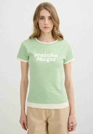 Joven mujer con cabello liso castaño claro lleva una camiseta de manga corta verde claro con el texto "Matcha Magic" y pantalones beige sobre un fondo blanco.