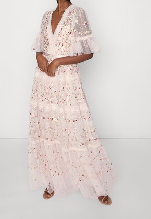 Femme portant une robe longue transparente rose pâle avec broderies florales et bordure en dentelle, debout les mains jointes devant.