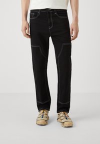 Svart denimbukser med hvite sømmer, med frontlommer og rett ben-designet. Brukt med beige sko med fargede aksenter.