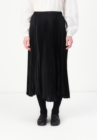Melna plisētā svārki ar elastīgu jostu, plūstoša auduma, garums līdz pusei teļiem, kombinēti ar melnajām zeķbiksēm un slip-on apaviem.