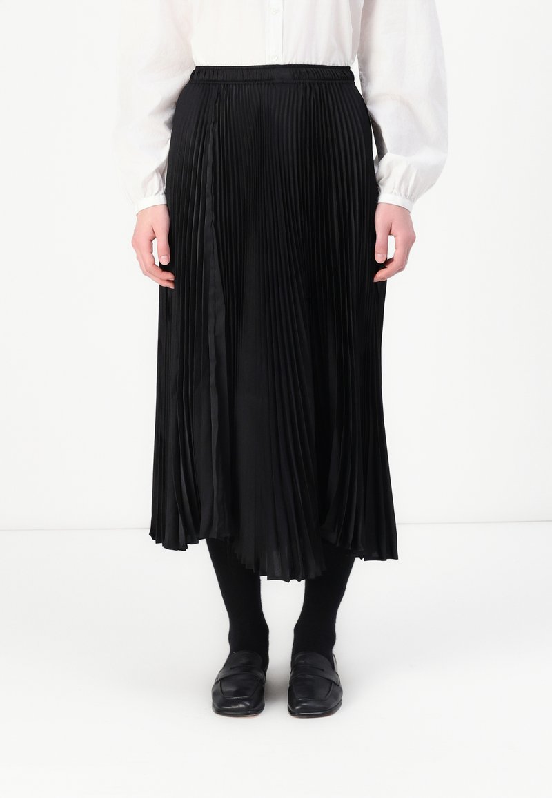 Fustă plisată neagră cu talie elastică, material fluid, lungime până la jumătatea gambei, purtată cu ciorapi negri și pantofi slip-on.